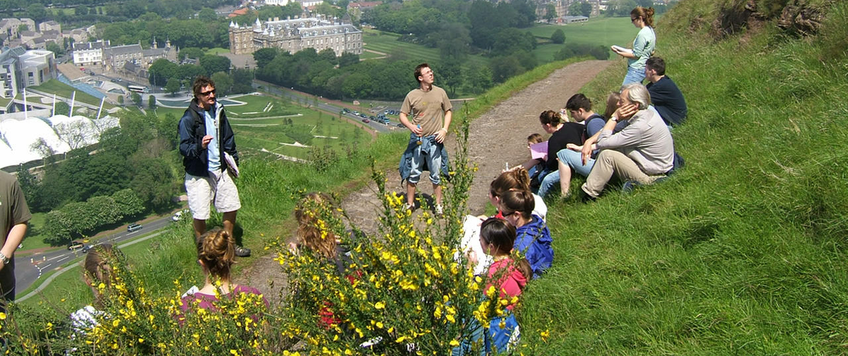 A group of students is gathered on a grassy Scottish hillside, with some sitting and others standing. The group appears to be in the middle of an outdoor lecture or discussion. In the background, a scenic view of a city with historic buildings, lush greenery, and modern structures is visible, likely showcasing a blend of urban and natural landscapes. Bright yellow wildflowers are in the foreground, adding color to the scene. 